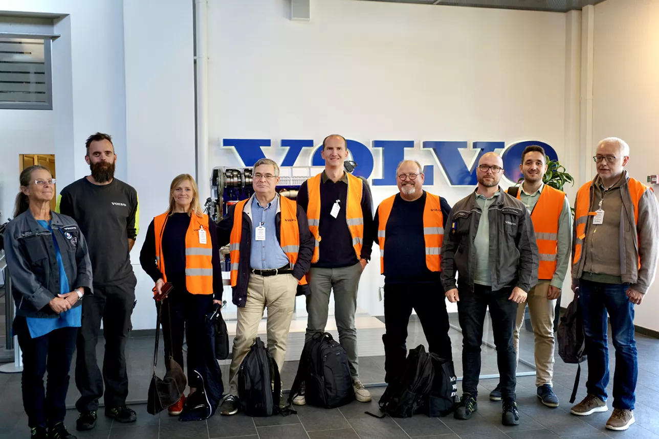 Photo of a group of people wearing orange vests, high visibility clothing, standing in front of a logotype.